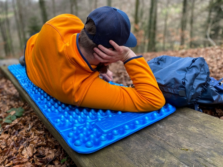 A person in an orange pullover and blue hat lounges on a blue Exped FlexMat sleeping pad