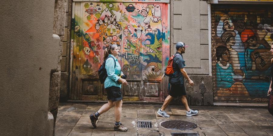 Two people walk along a city street in front of graffiti.