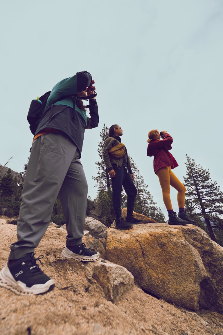 Three people stand on a rocky outcropping looking off at the distance. Two are using binoculars.