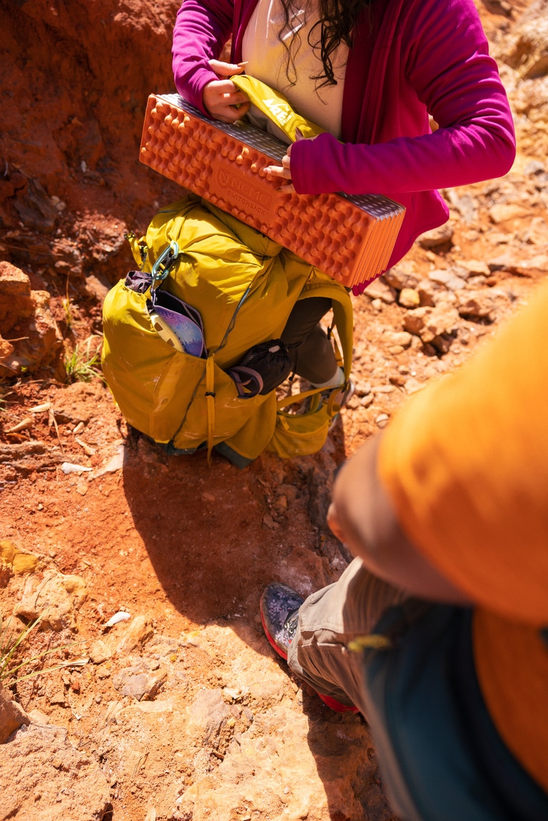 Two people on red dirt, one is securing a NEMO Switchback pad to their hiking pack