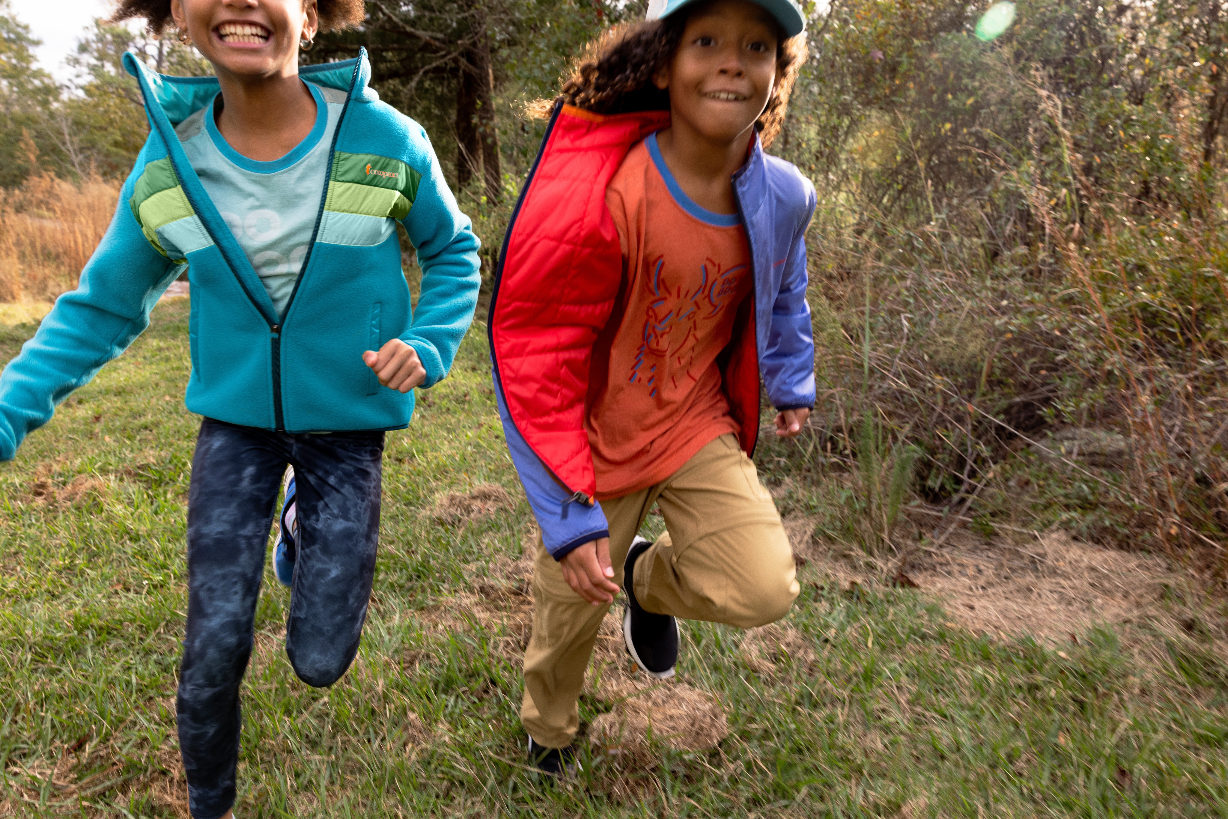 Two kids in hiking clothes running through a field.