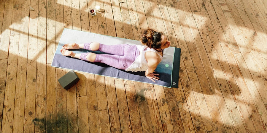 An adult wearing lightweight fitness attire does a yoga pose on a mat on a wooden floor, a foam block nearby