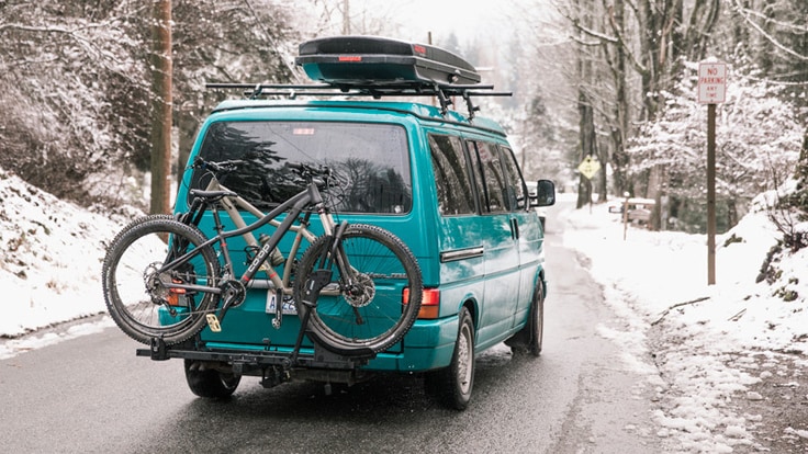 A van carrying bikes on a hitch rack and a cargo box on the roof rack drives down a snowy road