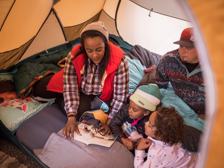 A woman reads a book to two kids inside a tent.