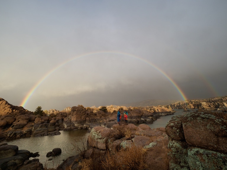 Two people standing on rocks underneath a rainbow in the desert.