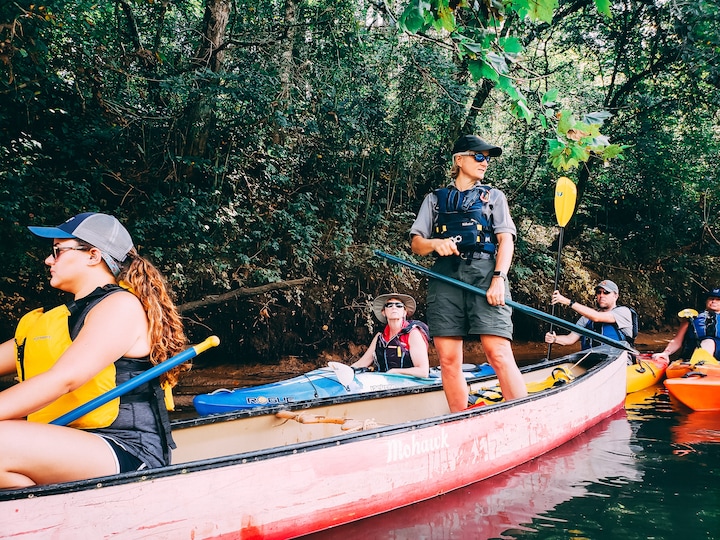 A guide stands on a canoe while other paddle in kayaks down a river