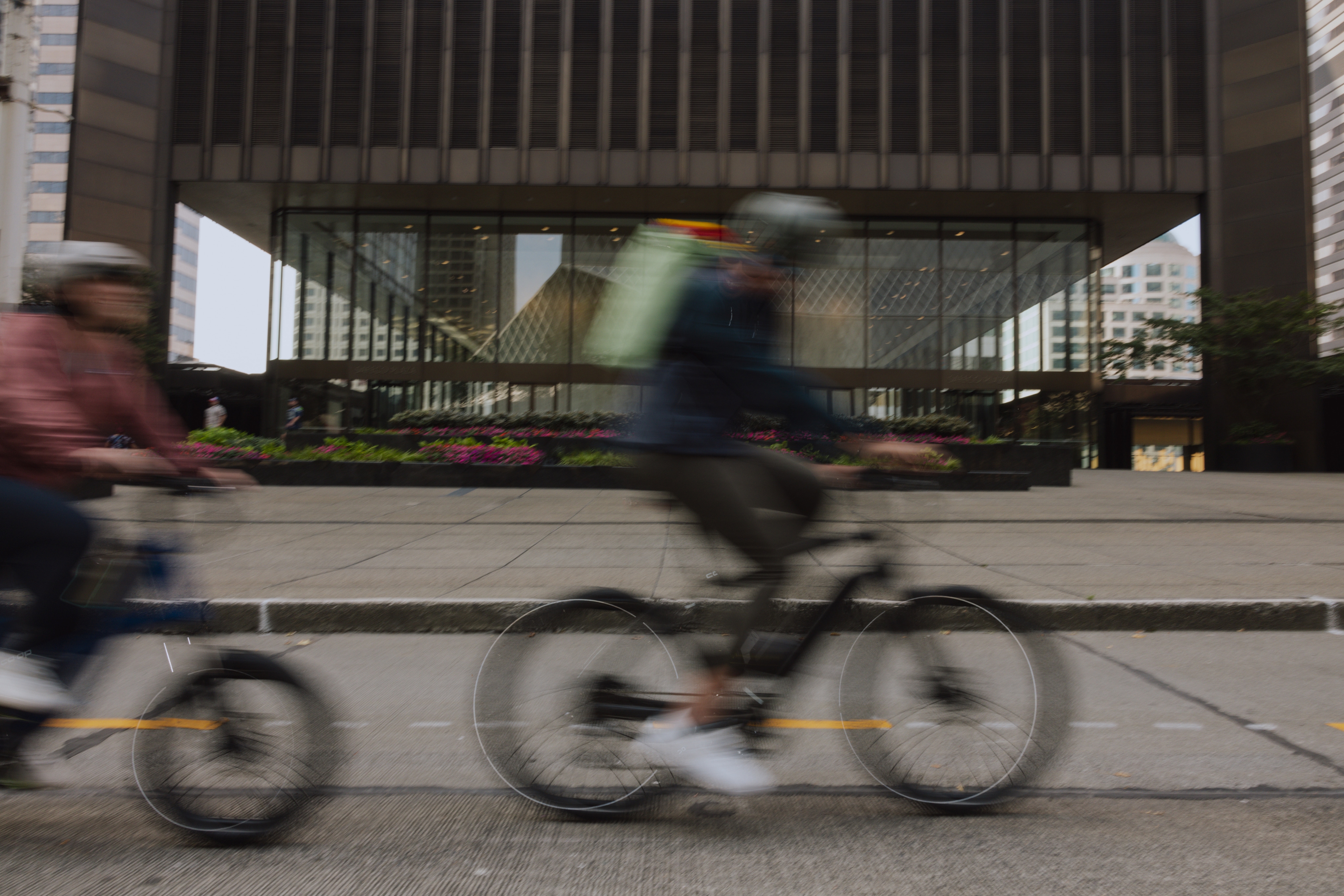 Two people pedal e-bikes along a road in front of a building