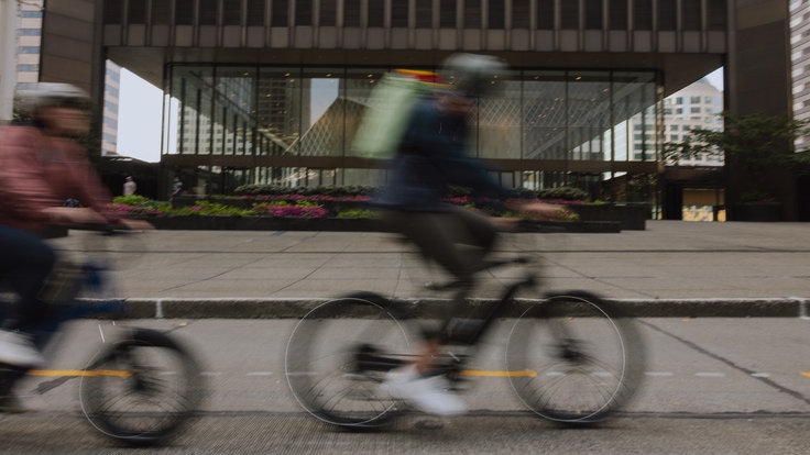 Two people pedal e-bikes along a road in front of a building