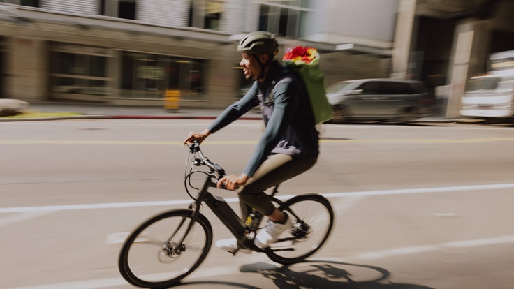 A person carrying flowers in a backpack pedals an electric bike through town