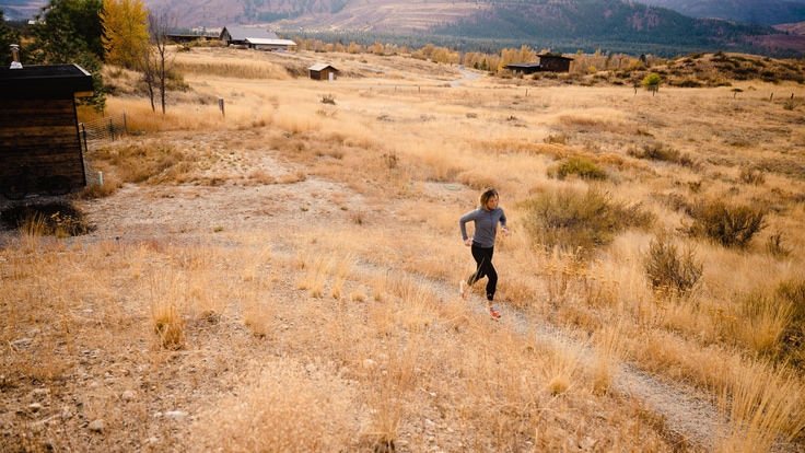 A runner traverses a trail with a large mountain range behind her and a small cabin to her right.