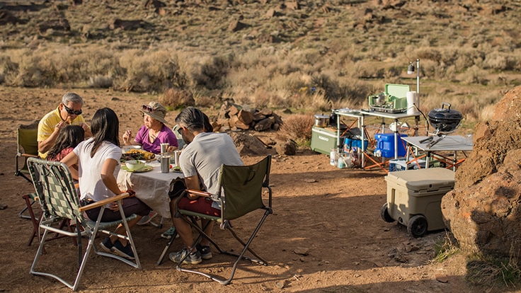 A family eats a meal at a campsite, near a homemade camp kitchen setup.