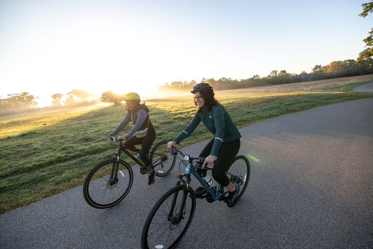 Two cyclists ride on a paved road at sunrise.