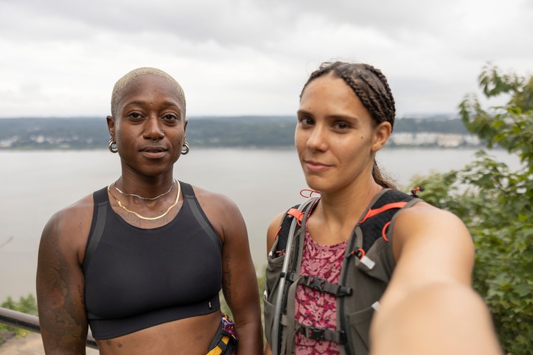 Two people, selfie view: one, with braids pulled back into a pony tail and wearing a hydration vest, holds the camera; the other, standing next to them, has a shaved head and wears a black sports bra. 