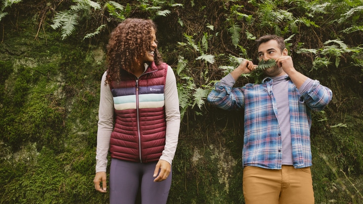 A woman and a man stand in front of a green hedge. They are both wearing base layer tops.