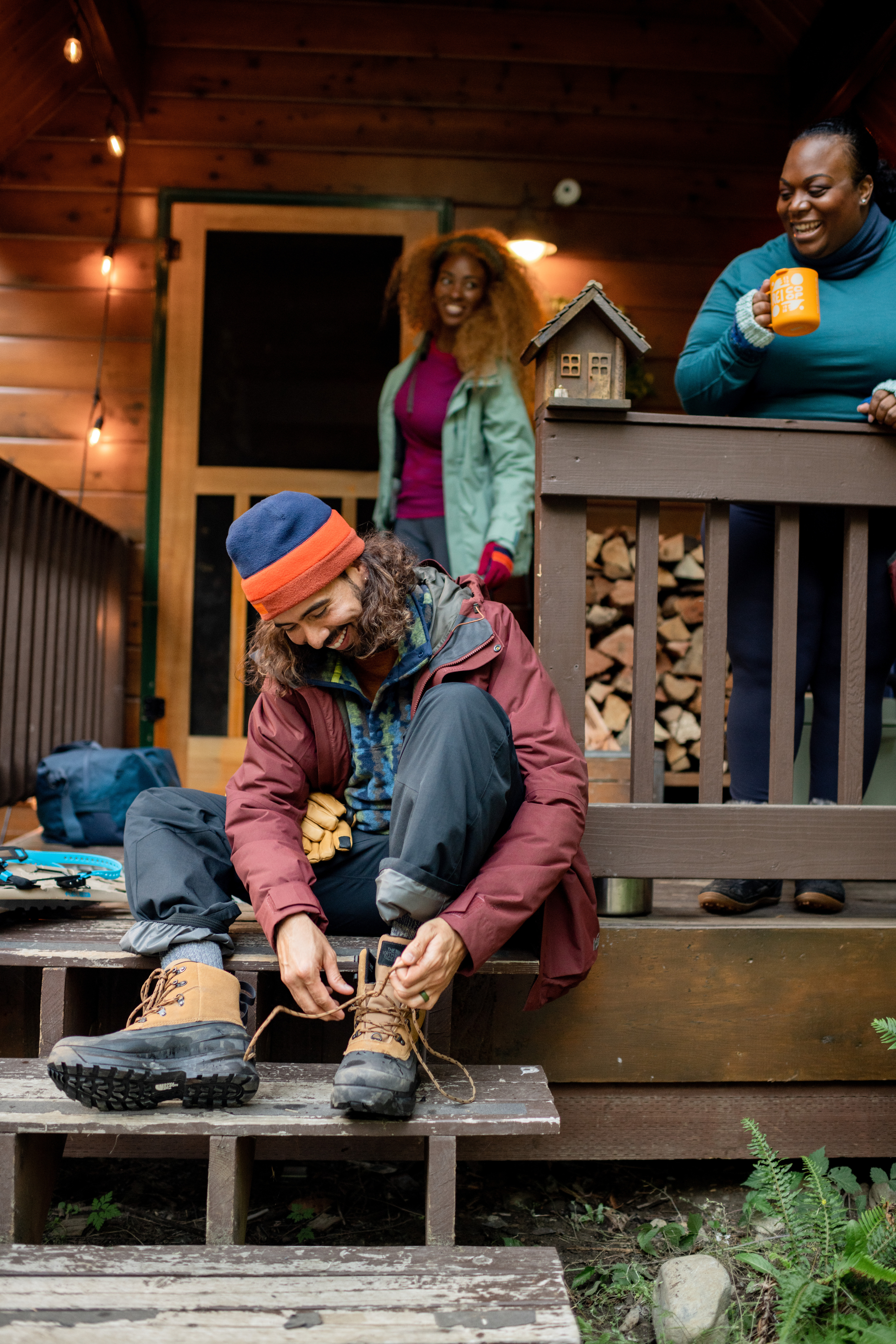 Friends spending time on cabin porch drinking from a mug while one ties up his boots