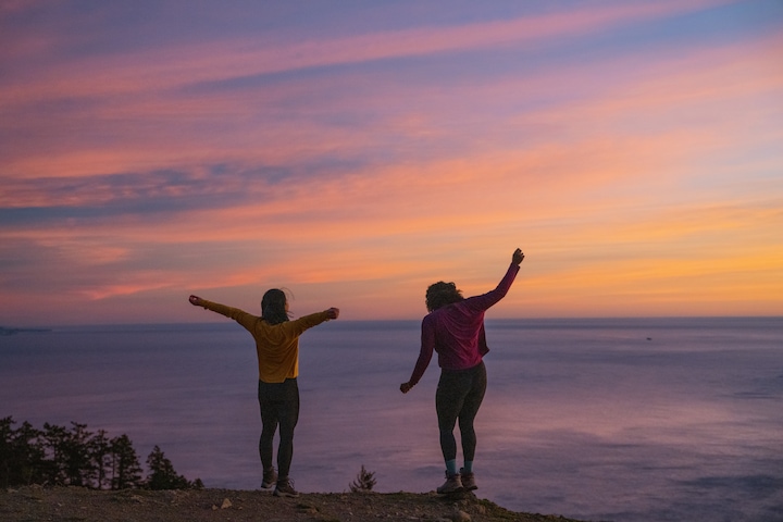 two hikers celebrating reaching the top of their hike overlooking a cloudy sunset