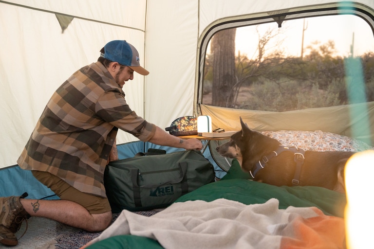 A person zips up a duffel inside a tent next to a dog