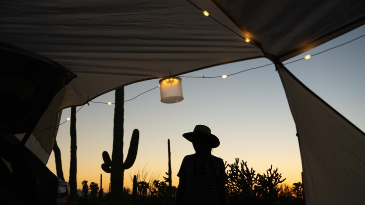 A person in shadow sitting at a desert campsite with string lights on the tent.