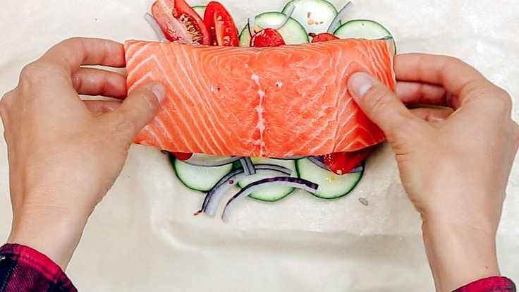 Two hands placing a salmon filet on top of cherry tomatoes and zucchini.