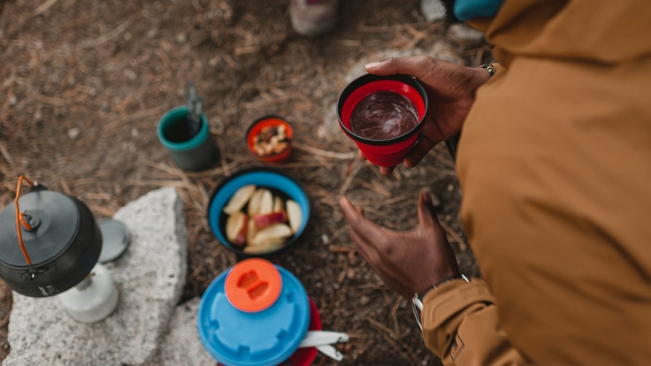 Person holds cup of coffee in the foreground. In the background there are snacks on the ground.