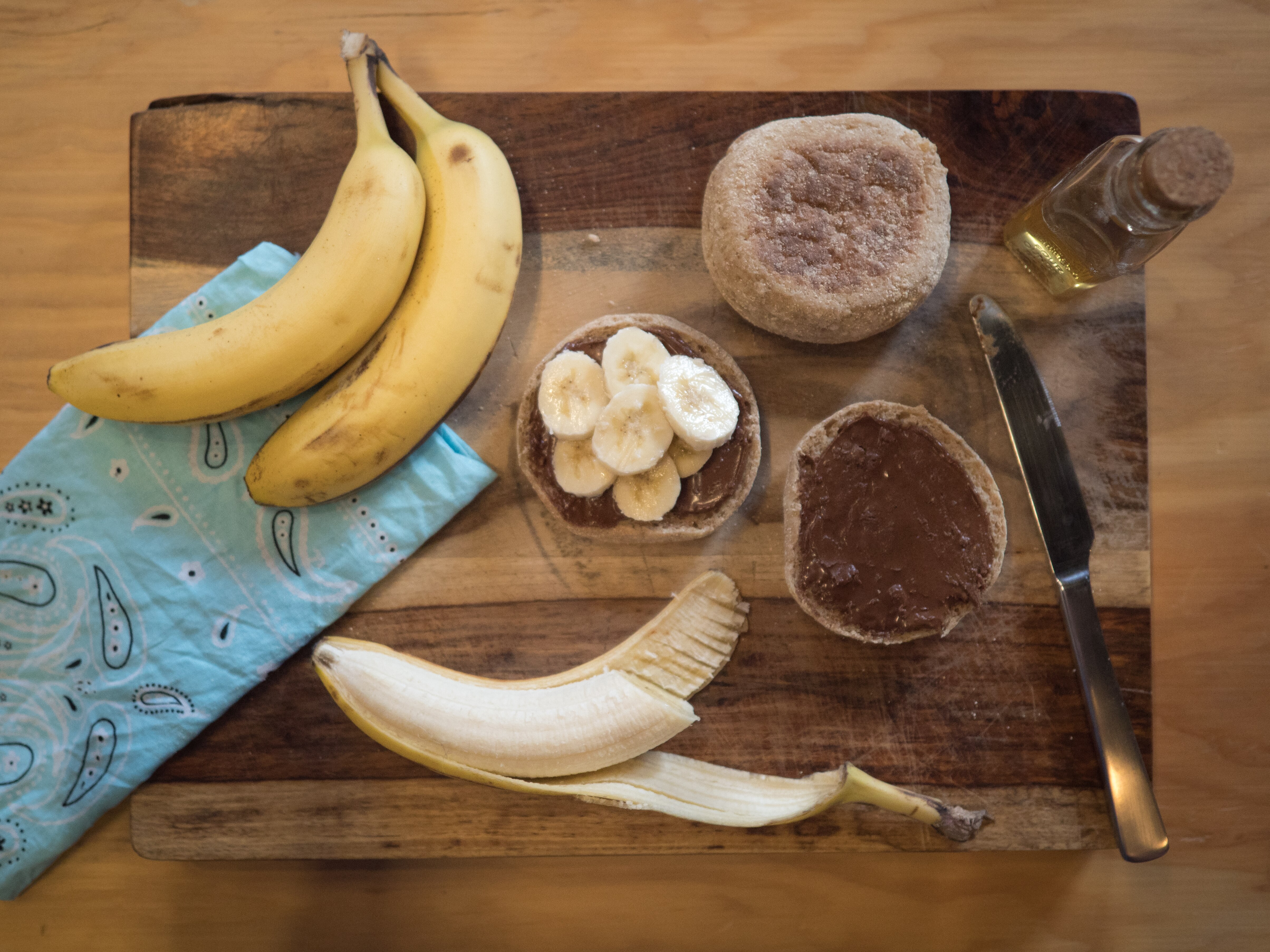 Banana, Nutella and English muffins are on a wooden cutting board.