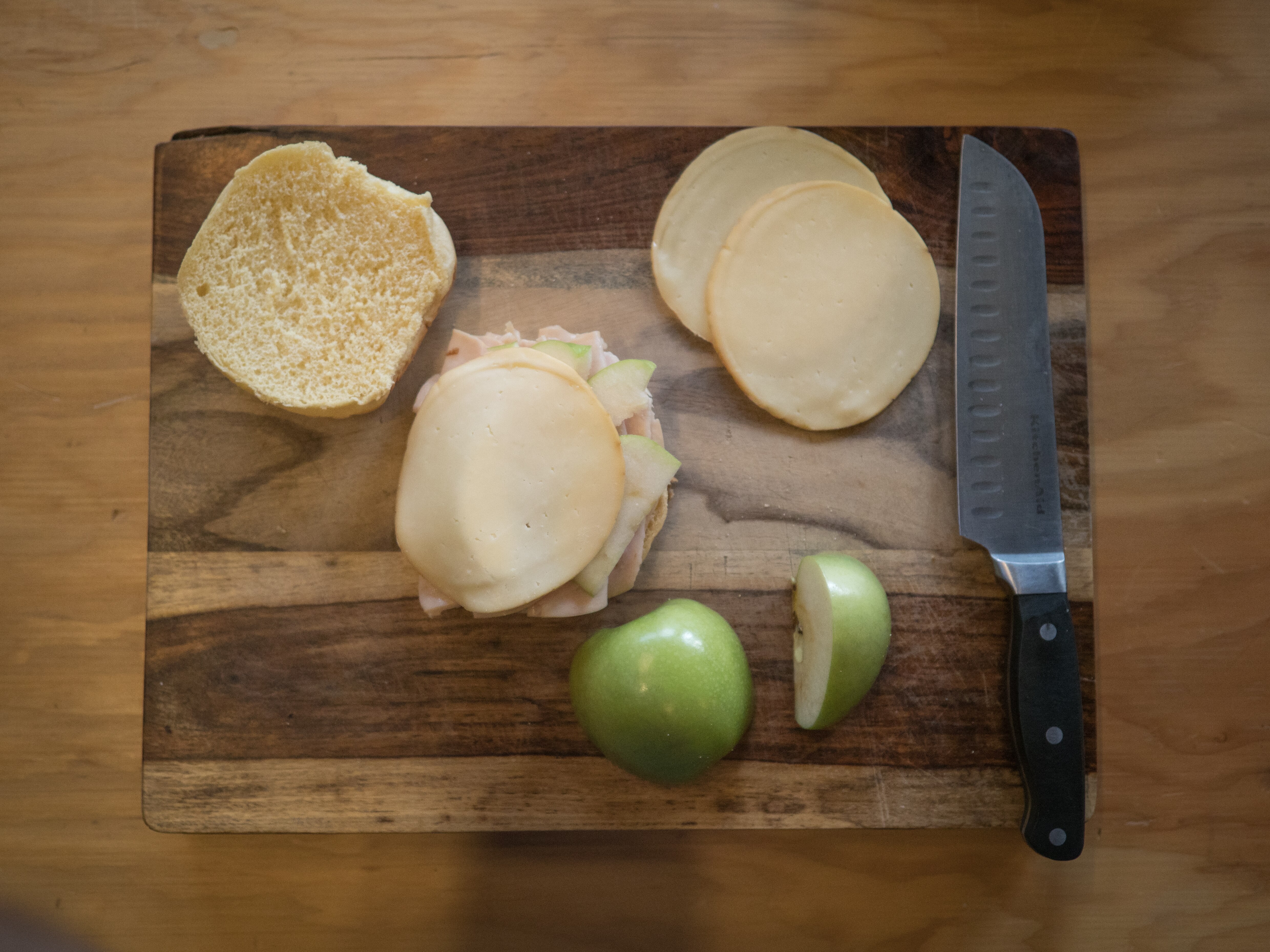 Bread, cheese, apple and knife are spread on a wooden cutting board.