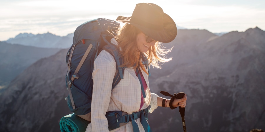 A long-haired hiker who is backlit by sunlight and wearing a pack, wide-brim sun hat and long-sleeve sun shirt