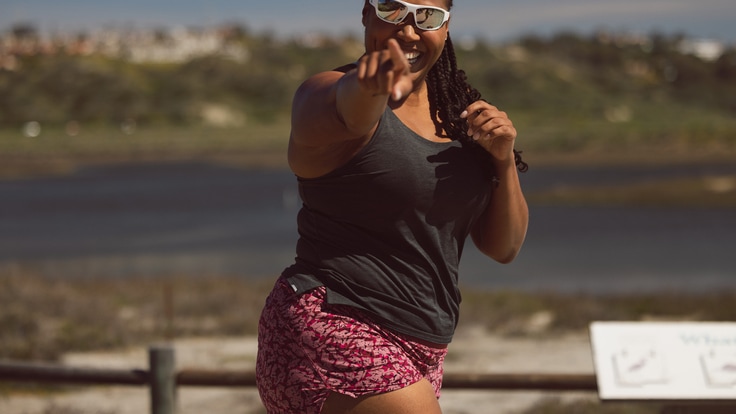 A person in pink patterned running shorts and a charcoal black running tank top and white sunglasses runs while pointing to the camera and smiling