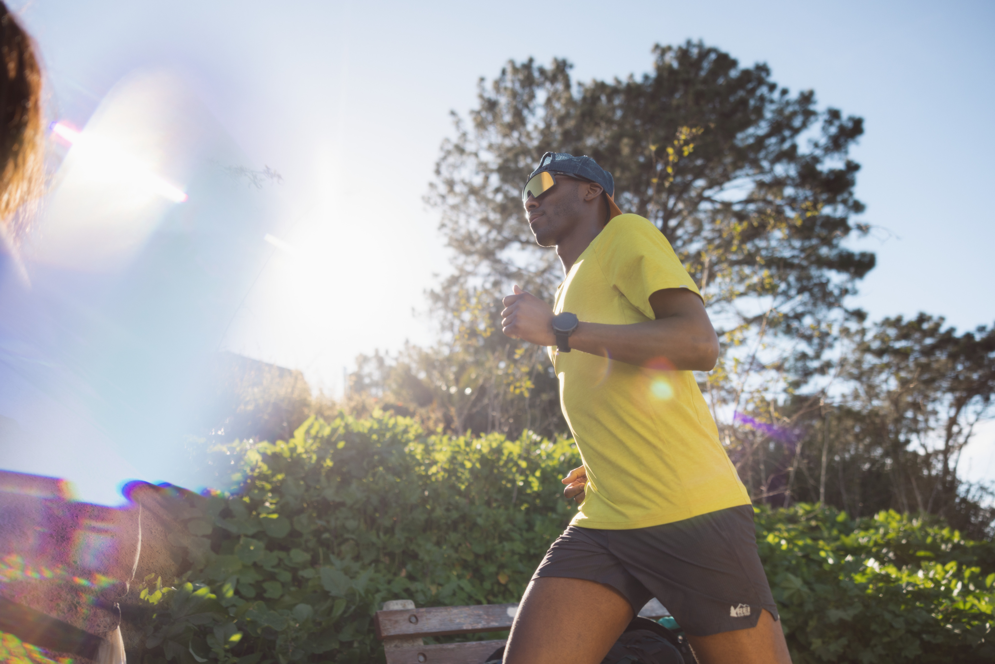 Man runs in the afternoon sun wearing grey shorts and a yellow shirt with his hat backwards and fun sunglasses