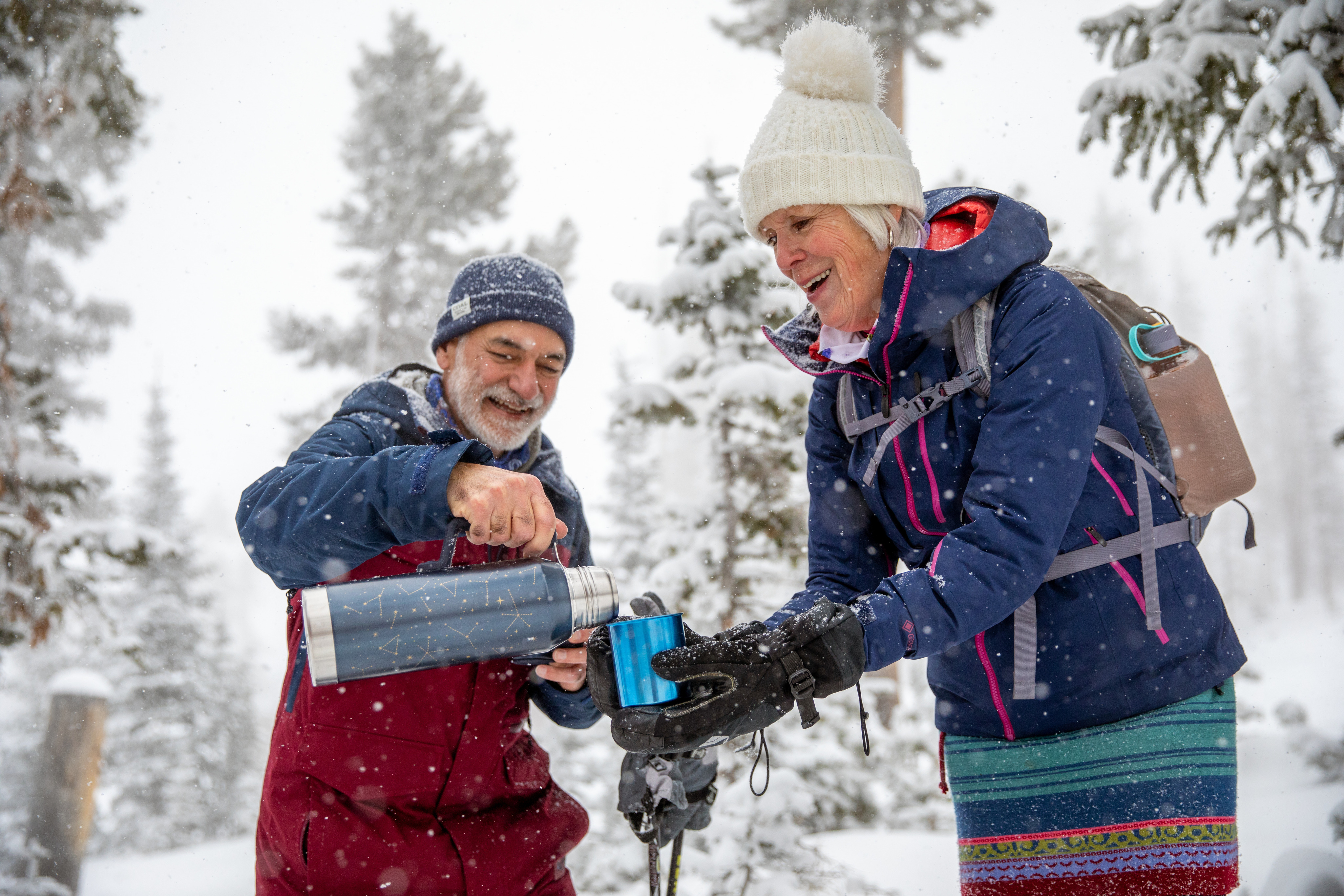 A man pours coffee from a thermos into a mug being held by a woman while snow rains down