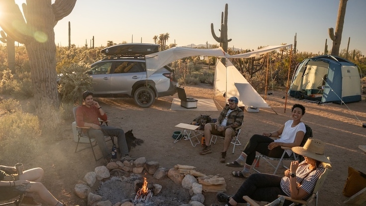 Campers sit around a fire in a desert setting. In the background is a car with a vehicle shelter attached and a tent.