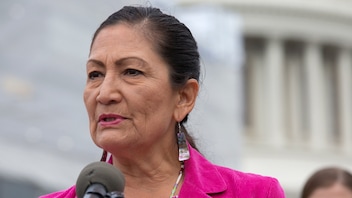 Secretary of the Department of the Interior, Deb Haaland, wearing a hot pink blazer, speaks into a microphone in front of a federal building.